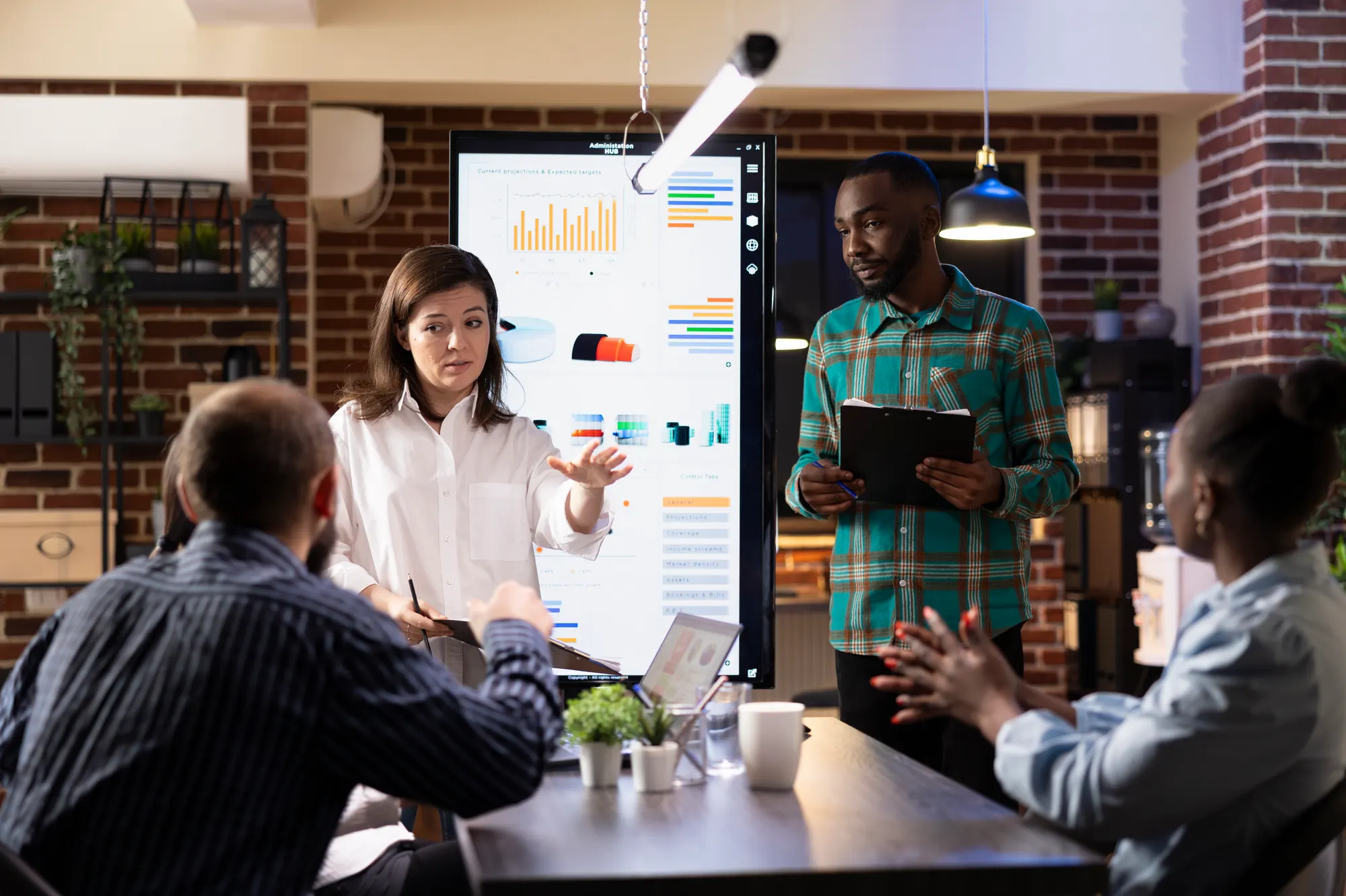 Two colleagues presenting growth charts on a large screen in a brick-walled office